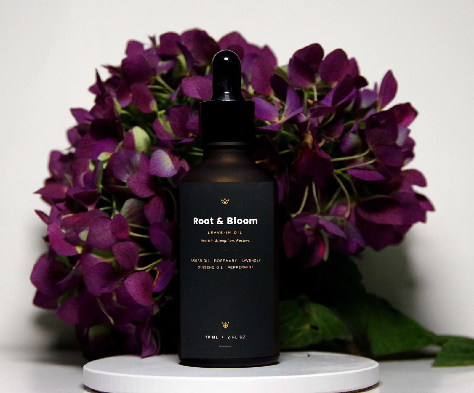 Root & Bloom Leave-In Oil dropper bottle between hydrangeas